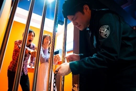 警察官のような制服の男性が牢屋のチェーンを操作しており、驚いた表情の男女が中にいる