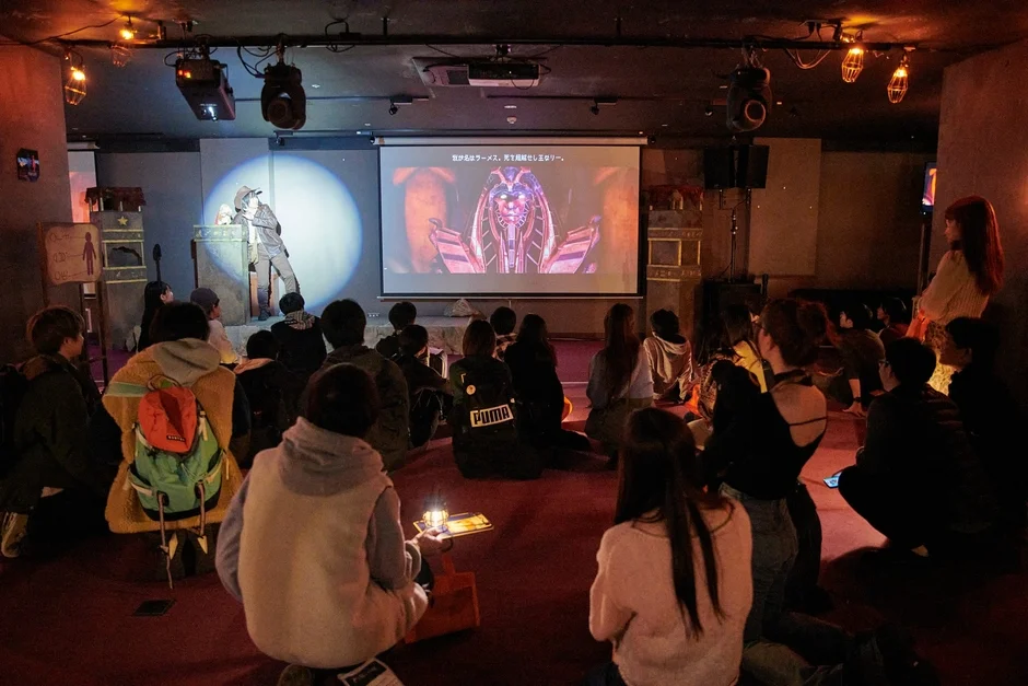 暗い部屋でスクリーンとステージ上のパフォーマーを見ているイベントの様子