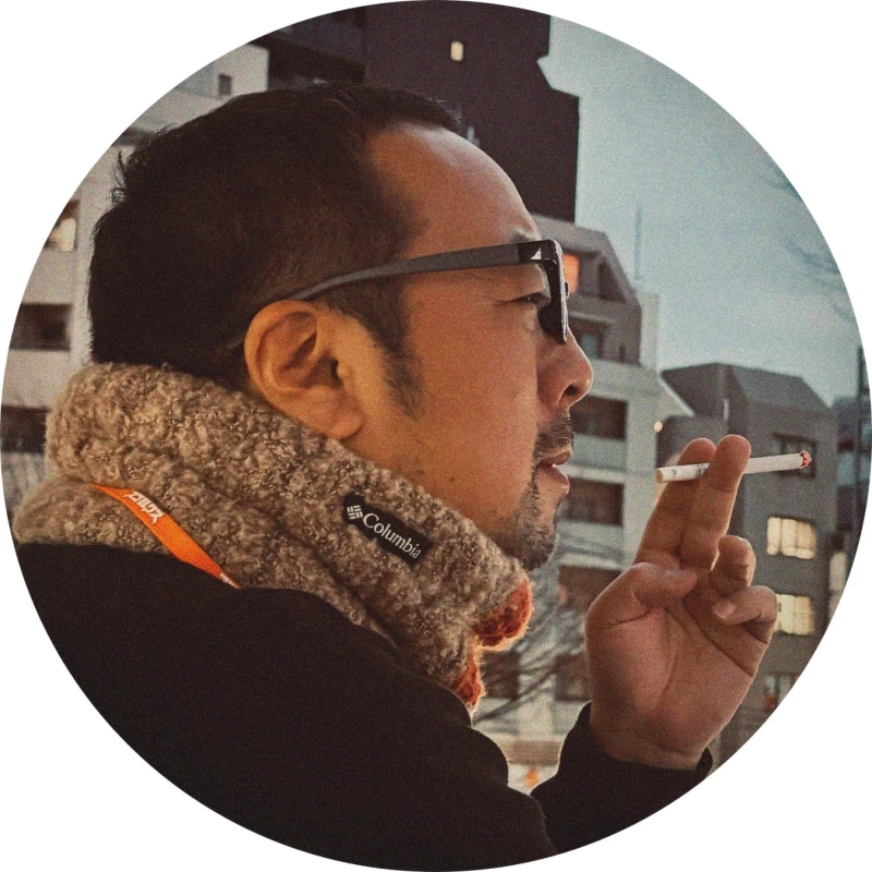 メガネとコロンビアのマフラーを着用した男性が、屋外でたばこを吸っている横顔の画像