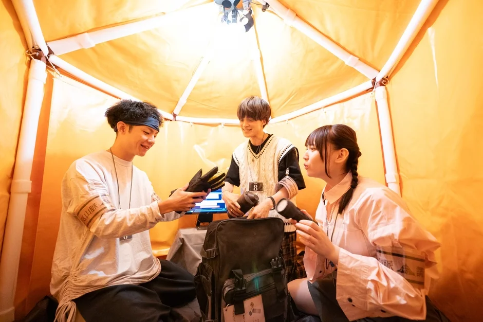 オレンジ色のテントのような空間に3人の若者が集まり、笑顔で会話をしています。