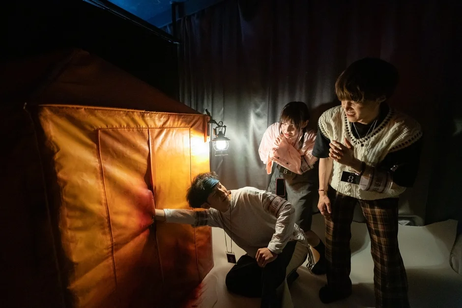 薄暗い室内で、3人の若者が緊張した面持ちでテントのような構造物に対峙しています。