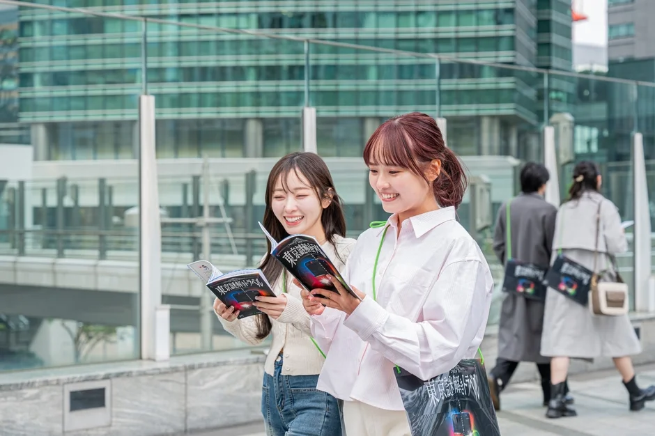 都会の街並みで、二人の若い女性が「東京謎解きトラベル」のパンフレットを笑顔で見ている