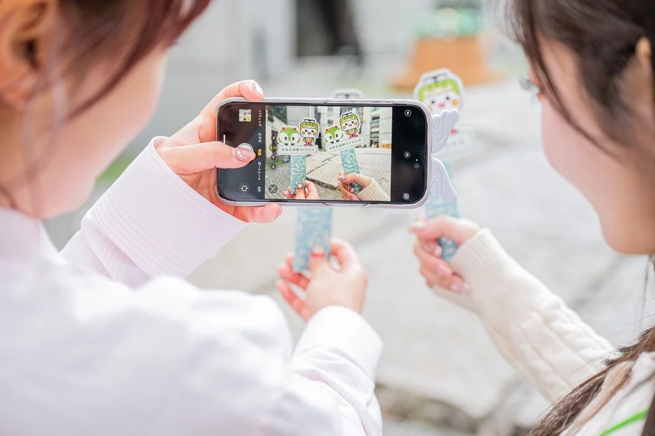 二人の女性がスマートフォンでキャラクターグッズを持ち、記念撮影をしている場面