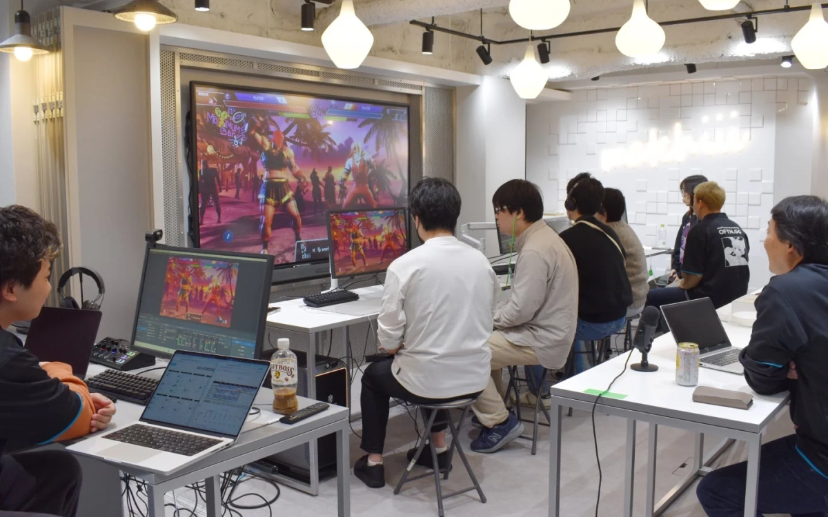 複数の男性がゲーミングスタジオのような場所で、対戦格闘ゲームの大画面を見ながらプレイしたり、パソコン作業をしている様子