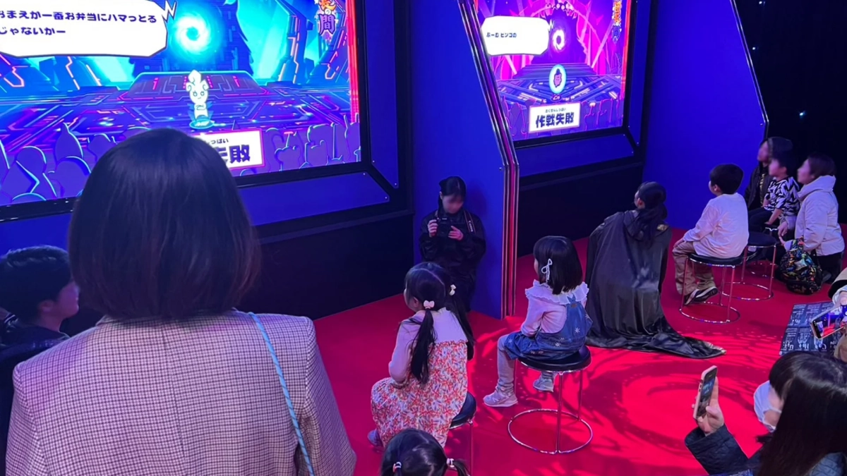複数の子供と大人が、明るいネオンカラーのゲーム画面が映る大型スクリーンを囲んで座っている様子