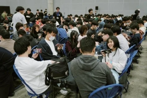 屋内の広い会場で、多くの若者がスマートフォンや携帯ゲーム機に没頭している様子