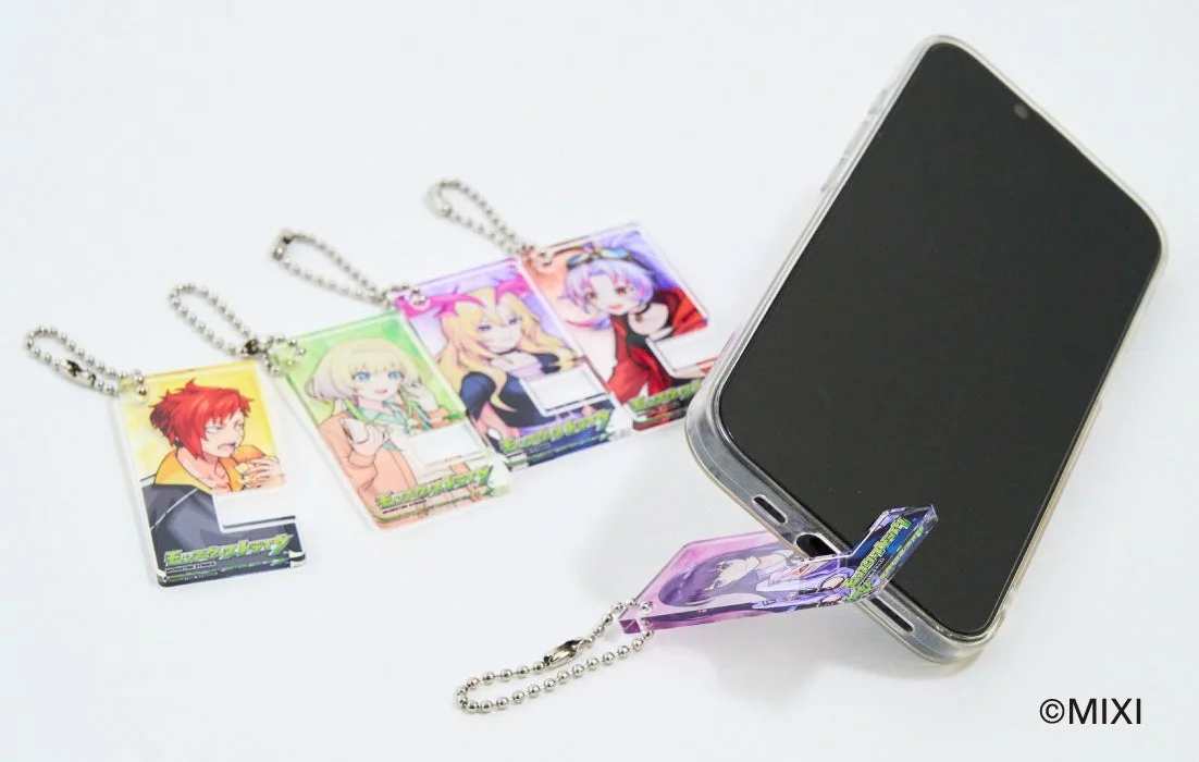 白色の背景に、アニメキャラクターが描かれた複数のアクリルキーホルダーと、キーホルダーをスタンドとして利用しているスマートフォンが写っています。