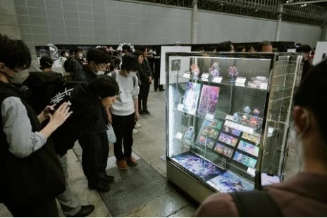 多くの人々が屋内イベント会場で、ガラスケースに陳列されたアニメやマンガ関連のグッズを見ている様子