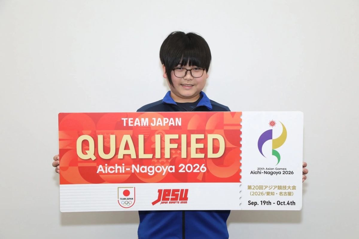 眼鏡をかけた人物が、2026年の愛知・名古屋で開催される第20回アジア競技大会への「TEAM JAPAN QUALIFIED」と書かれた大きなボードを持って笑顔を見せている