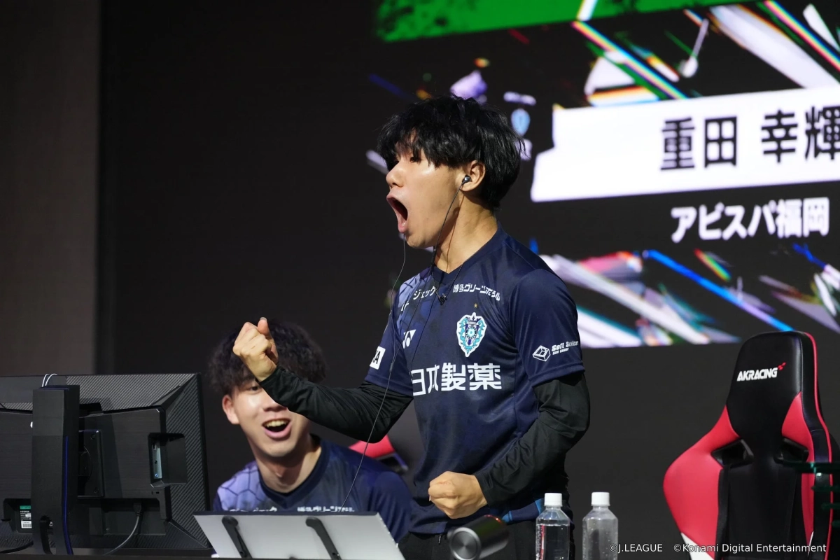 eスポーツの試合中に、青いユニフォームを着た男性選手が拳を握りしめ、口を大きく開けて感情を爆発させている瞬間。背景には「重田 幸輝」「アビスパ福岡」の文字が見え、勝利や熱戦の興奮が伝わってくる。