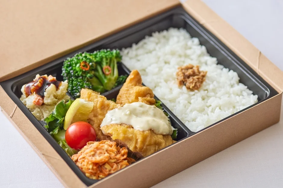 紙製の蓋付き容器に入った、魚フライ、ポテトサラダ、ブロッコリー、ご飯などが彩り豊かに詰められた和風のお弁当です。様々な種類のおかずがバランス良く配置され、美味しそうな一食を演出しています。