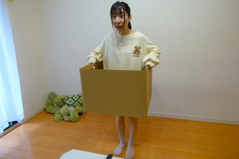 白いスウェットを着た若い女性が、大きな段ボール箱に入って部屋に立っています