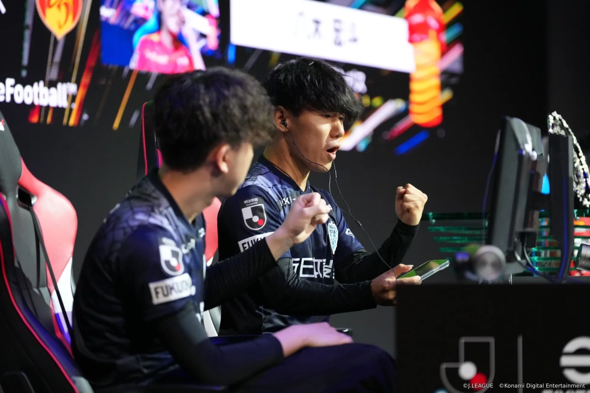 eスポーツの試合中に、Jリーグのユニフォームを着た選手がモニターに向かい、両手を握りしめ興奮している様子。隣にも別の選手が座っており、真剣な表情でゲームに集中する様子が伝わる。