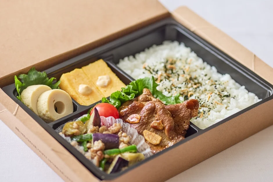 茶色い紙製の箱に入った和風のお弁当です。ふりかけのかかったご飯と、豚肉の生姜焼き、茄子や卵焼き、ちくわなどの彩り豊かな副菜がバランス良く詰められています。