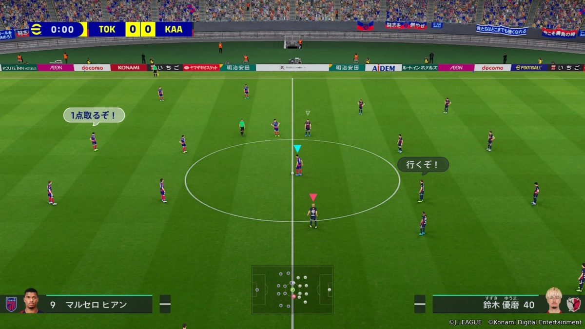 サッカーのビデオゲーム「eFootball」の試合画面。TOK対KAAのスコアは0-0。選手たちがフィールドに配置され、マルセロ・ヒアンと鈴木優磨の情報が表示されている。スタジアムには観客とスポンサー広告が見える。