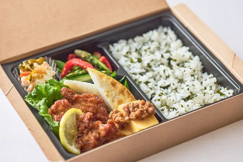 茶色い紙箱に詰められた和風のお弁当です。メインのから揚げをはじめ、卵焼き、かまぼこ、ポテトサラダ、野菜など彩り豊かでおかずが豊富。ふりかけのかかったご飯も入っており、ランチやテイクアウトにぴったりの一品です。