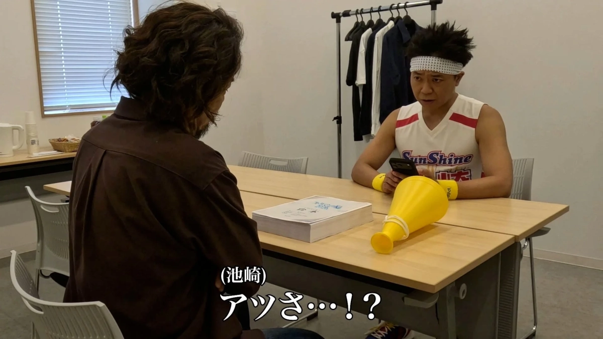 会議室で二人の男性が向かい合って座っています。一人はスポーツウェア姿で驚いた表情をしており、テーブルにはメガホンとスマートフォンが置かれています。