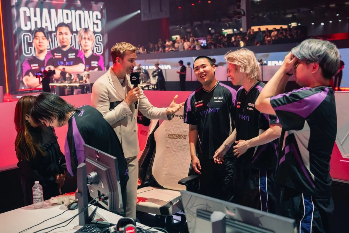 eスポーツ大会のステージで、マイクを持ったインタビュアーが黒と紫のユニフォームを着た選手たちに話しかけている様子
