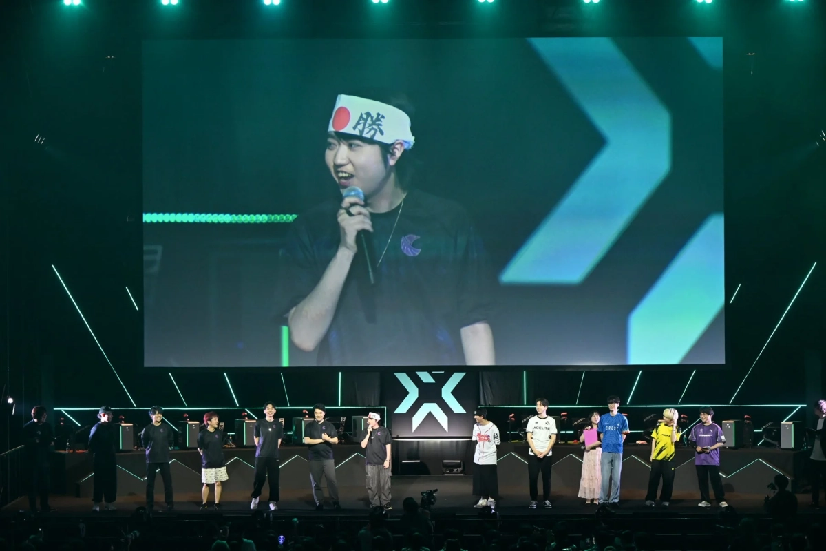eスポーツイベントのステージで、大画面に「勝」のハチマキを巻いた人物がマイクを手に話しています