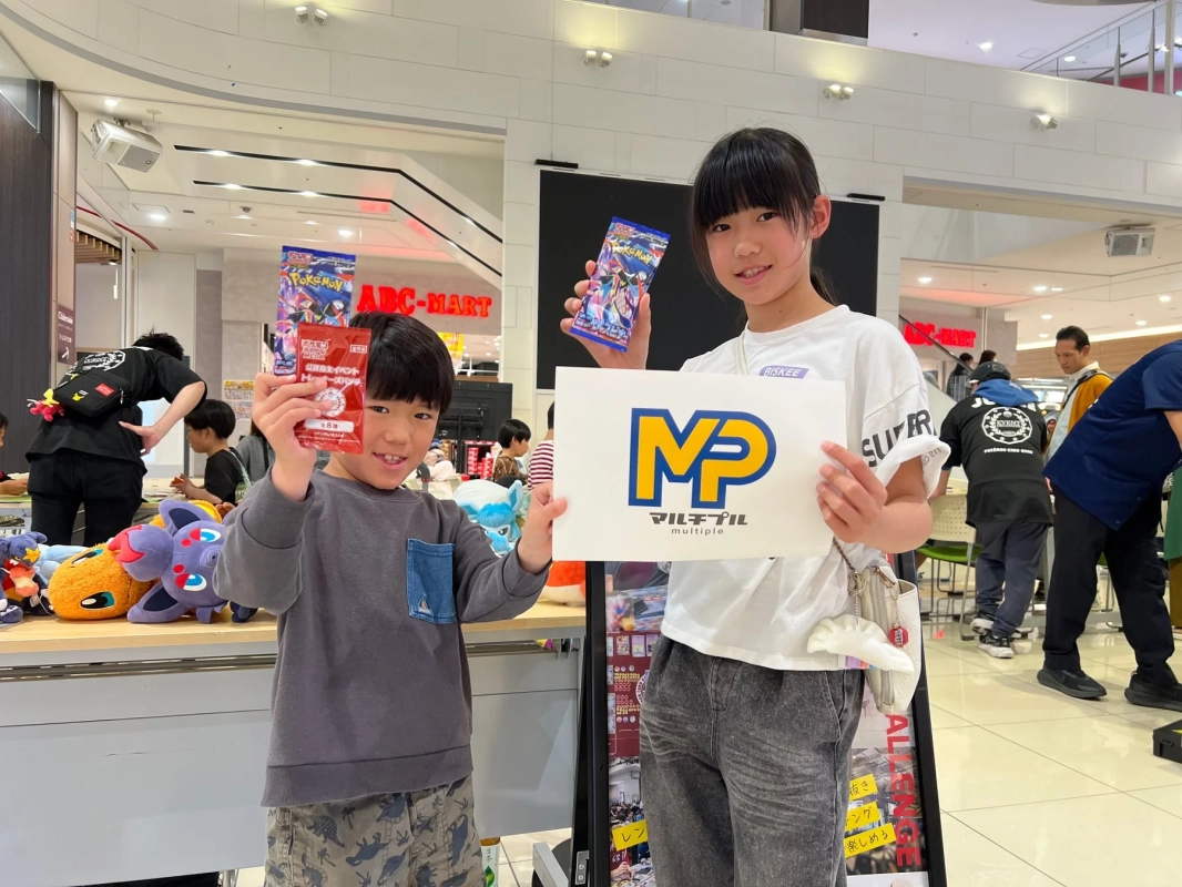 男の子と女の子がポケモンカードパックとロゴ入りボードを持つ、イベントの様子