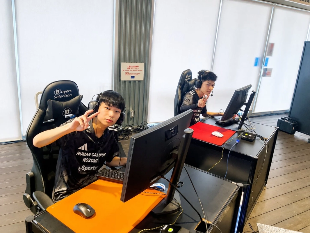 2人の若者がeスポーツイベント会場のような場所で、ゲーミングチェアに座りヘッドセットを装着してパソコンに向かっている。手前の男性はピースサインをしている。