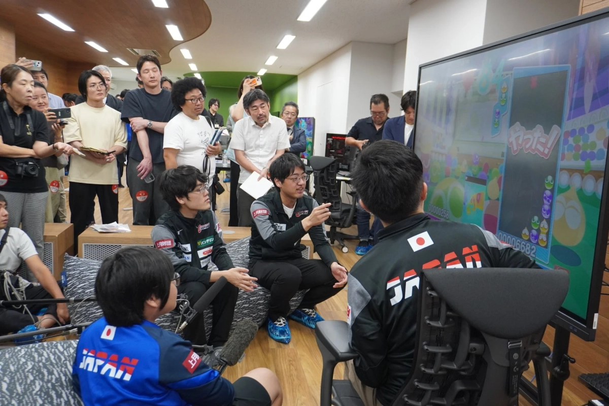 JAPANのロゴが入ったウェアを着た若者たちが、大型モニターに映し出されたゲーム画面を見ながら真剣に議論している様子