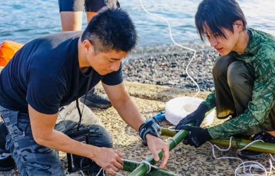 水辺で竹とロープを組み立てる男性二人