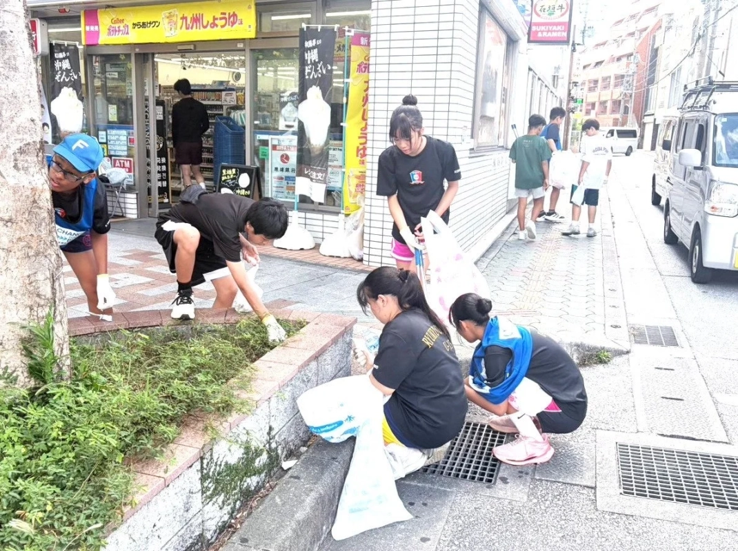 複数の若者たちが、日本の商店街の歩道や植え込みでゴミ拾いや清掃活動を行っている様子です。軍手をはめ、ゴミ袋を持ち、協力して街をきれいにしています。